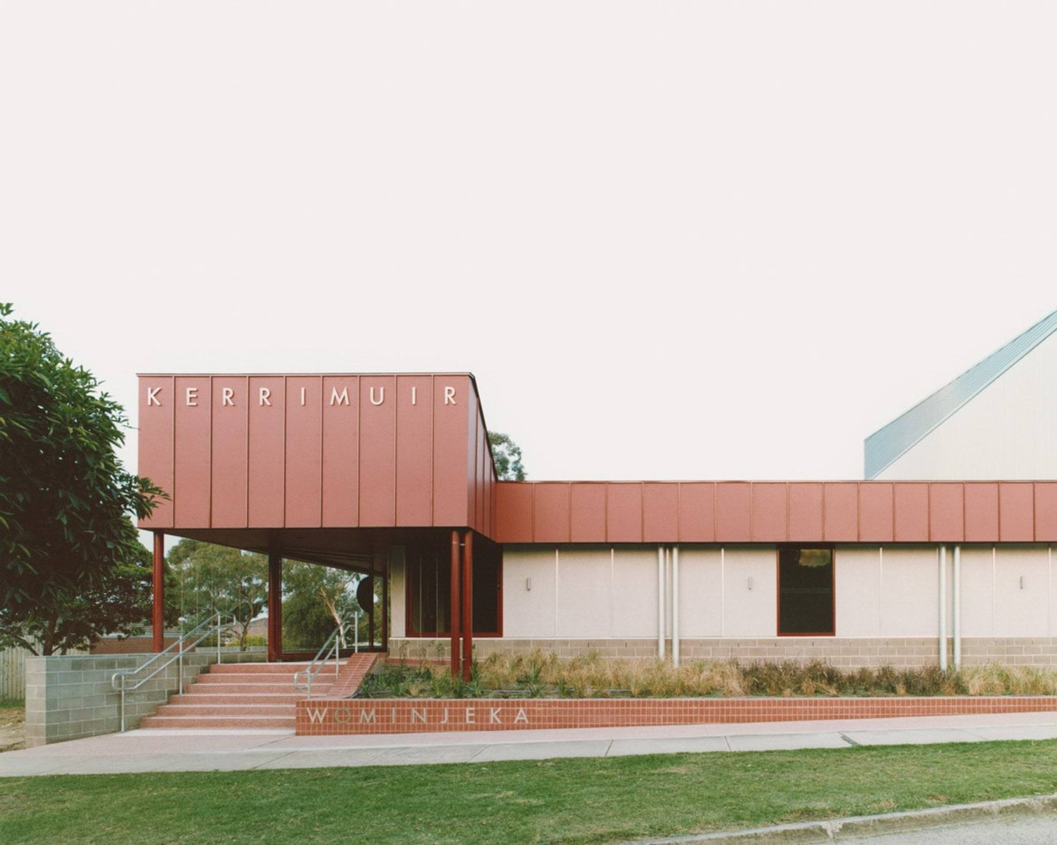 The front view of the school entrance with bold red cladding and “KERRIMUIR” signage above. Steps and landscaped garden beds lead to the doorway.