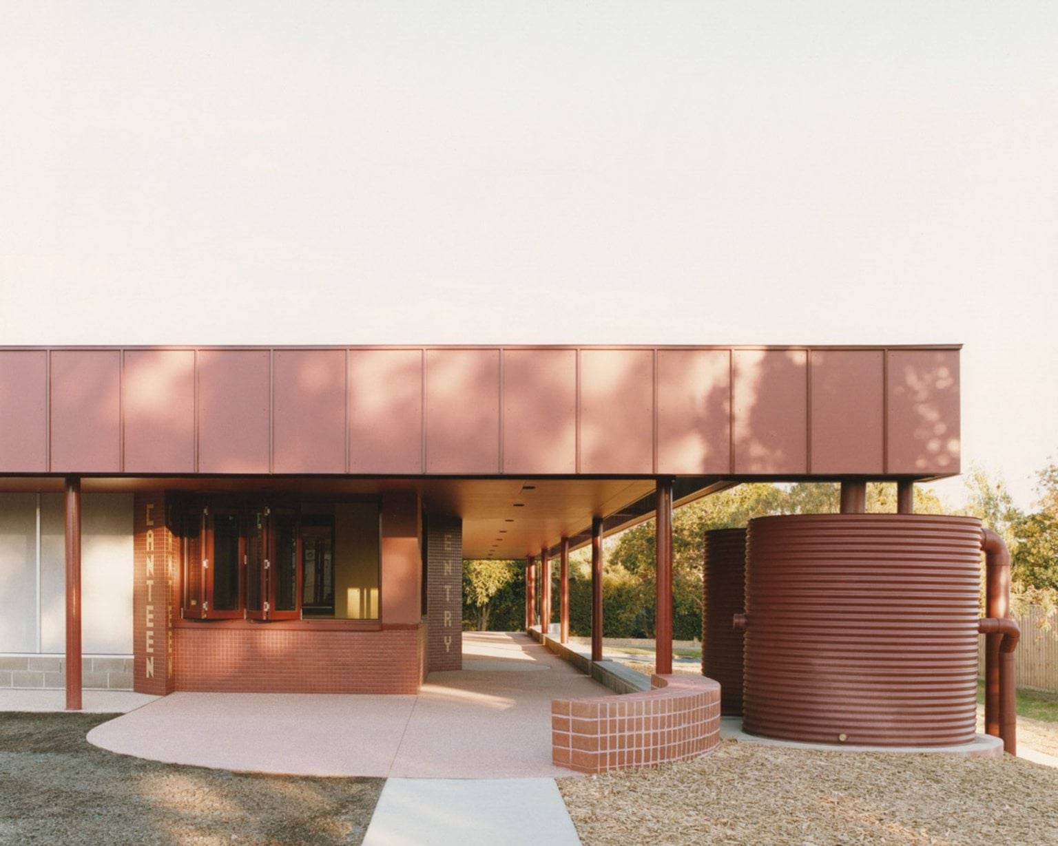 A covered outdoor area with red cladding and signage for “CANTEEN” and “ENTRY.” Two large cylindrical water tanks are positioned beside the building.