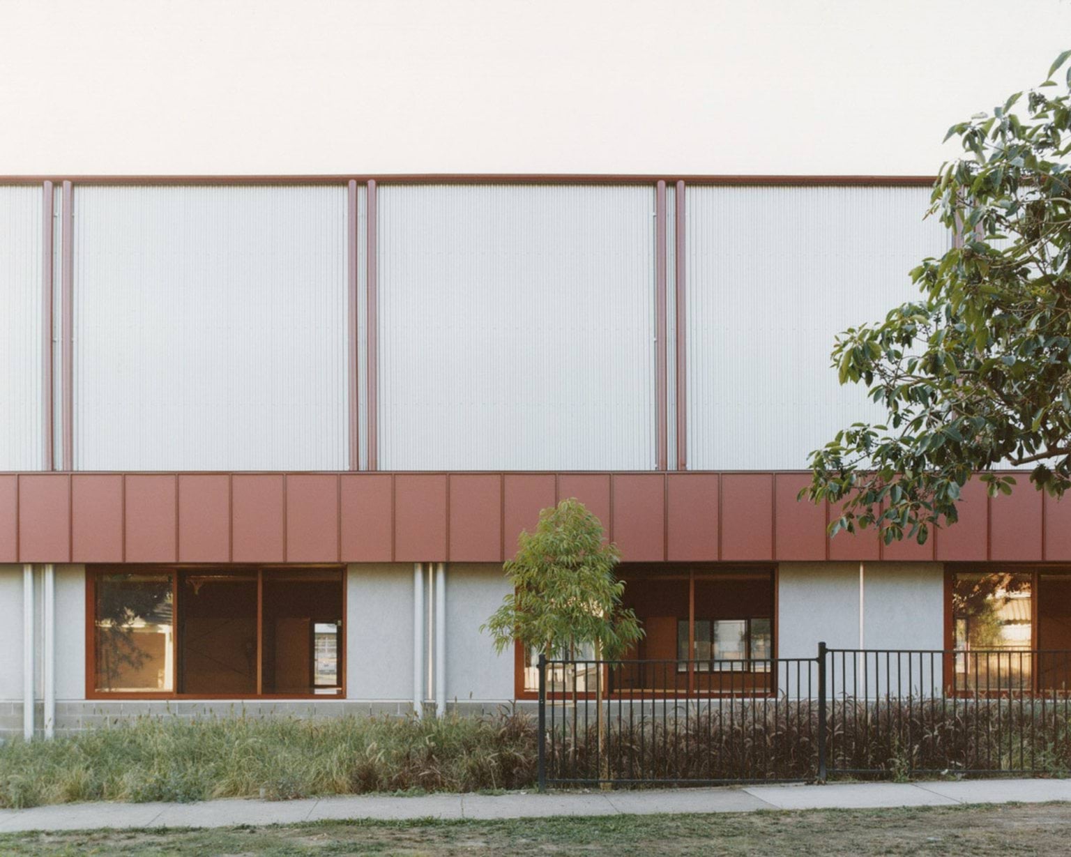 A side elevation of the school building with red cladding and translucent panels above grey walls. A black metal fence and young trees line the footpath.