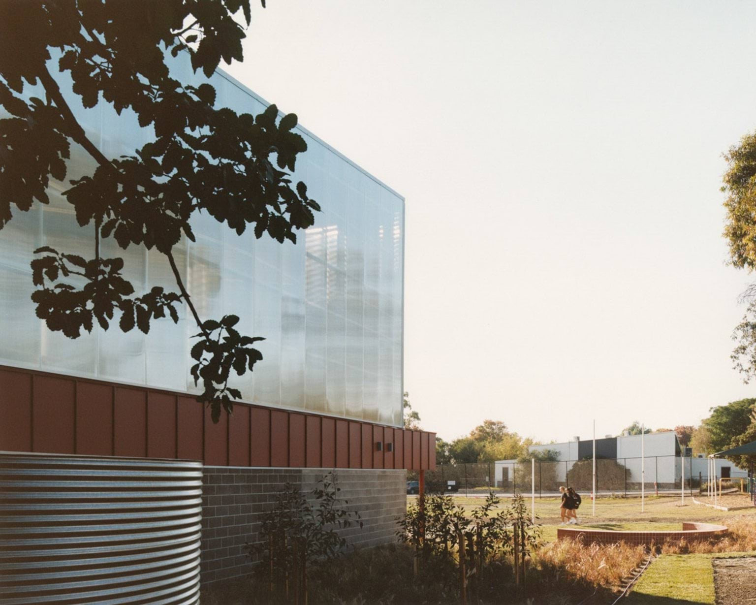 The exterior of a modern school building with translucent wall panels, red feature cladding, and grey brickwork. A landscaped area with young plants and a curved brick seating wall is in the foreground.