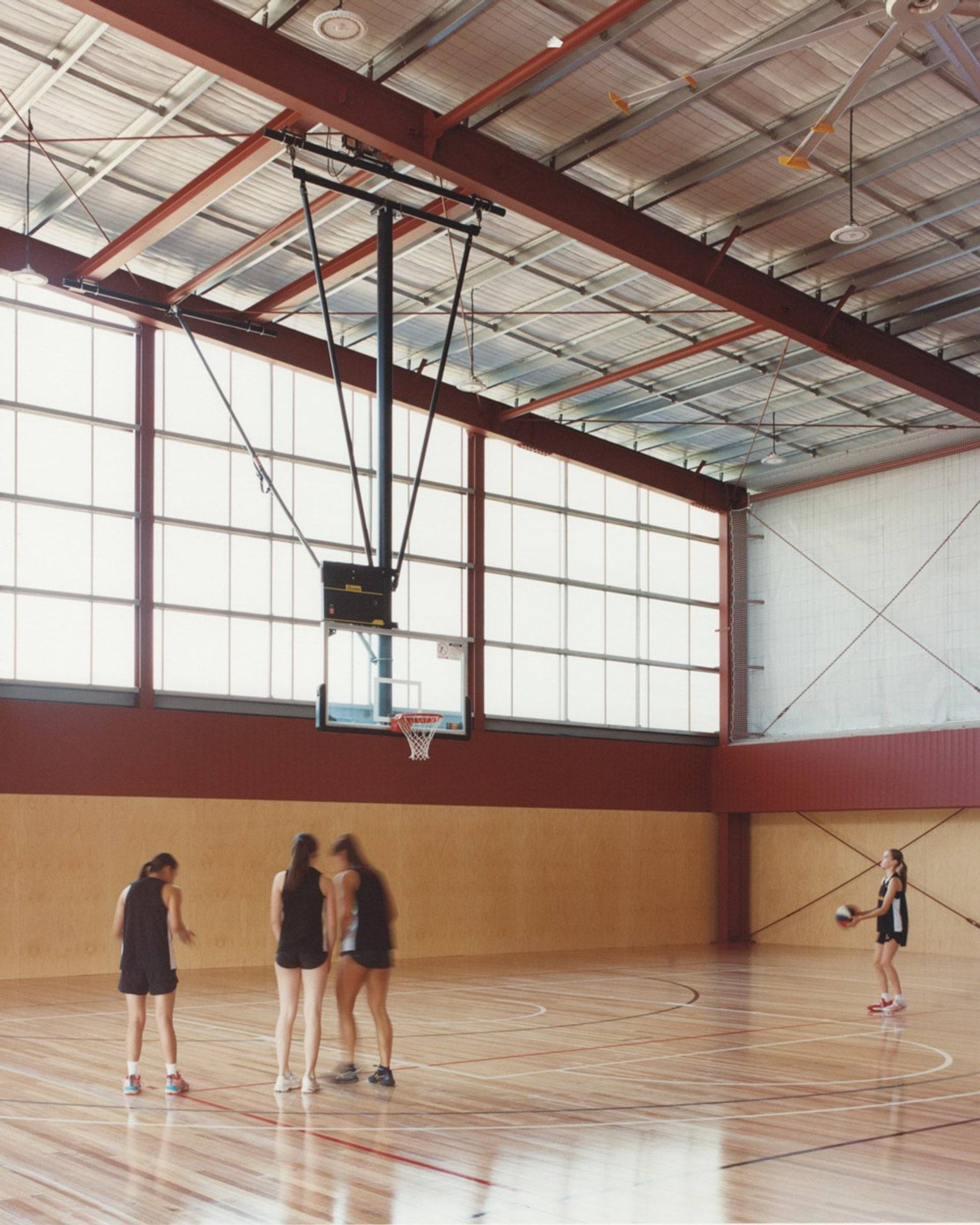 An indoor basketball court with timber flooring, high ceilings, and large windows. Four students stand near the hoop while another holds a basketball.