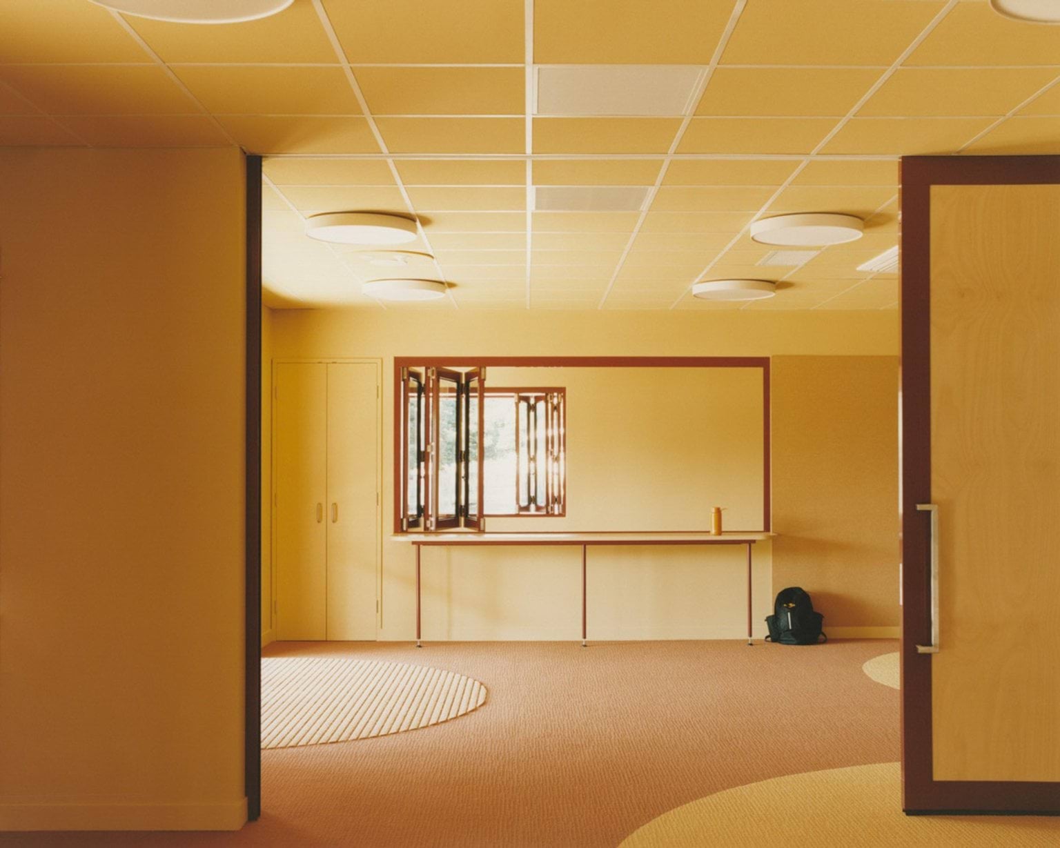 An indoor learning space with yellow walls, circular carpet inlays, and round ceiling lights. A timber-framed window and bench are visible at the far end.