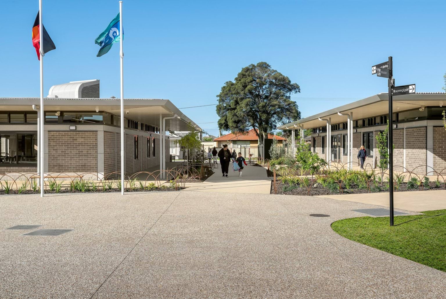 A landscaped courtyard with two flagpoles, garden beds, and paved walkways between modern brick school buildings.