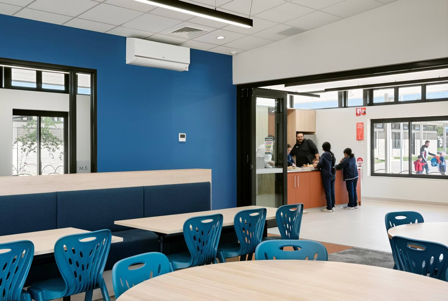 An indoor canteen space with blue walls, round tables, and blue chairs. A serving counter opens to the outdoor area through glass doors.