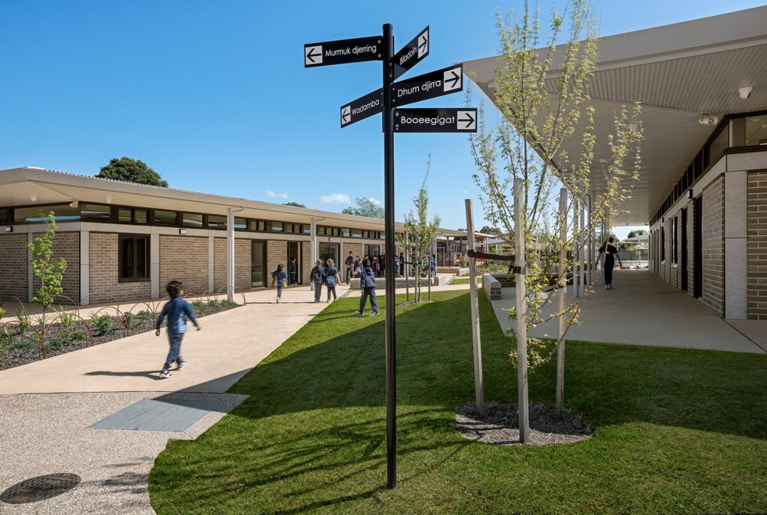 A landscaped school walkway with garden beds, young trees, and a black directional signpost. Students are walking between low-rise brick buildings with covered verandahs.