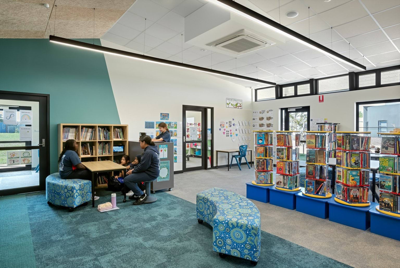 A school library with teal walls, patterned seating, and bookshelves filled with books. Round display units hold additional books in the centre.