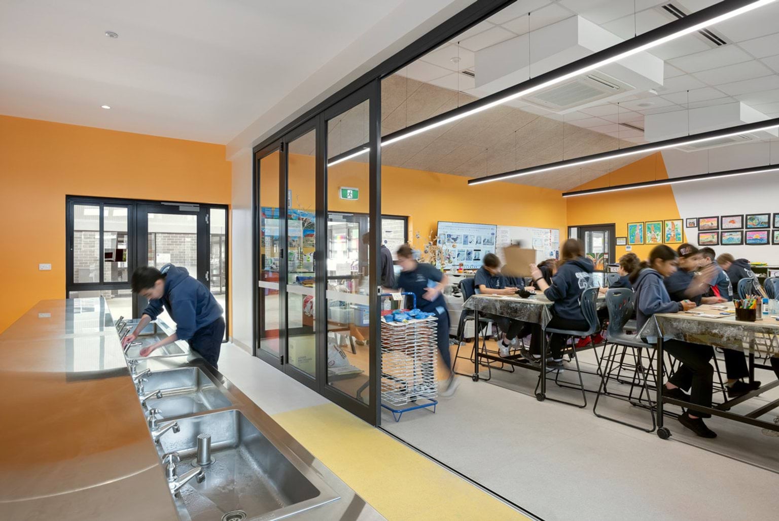 An art classroom with orange walls, stainless steel sinks along one side, and students working at tables with art supplies.