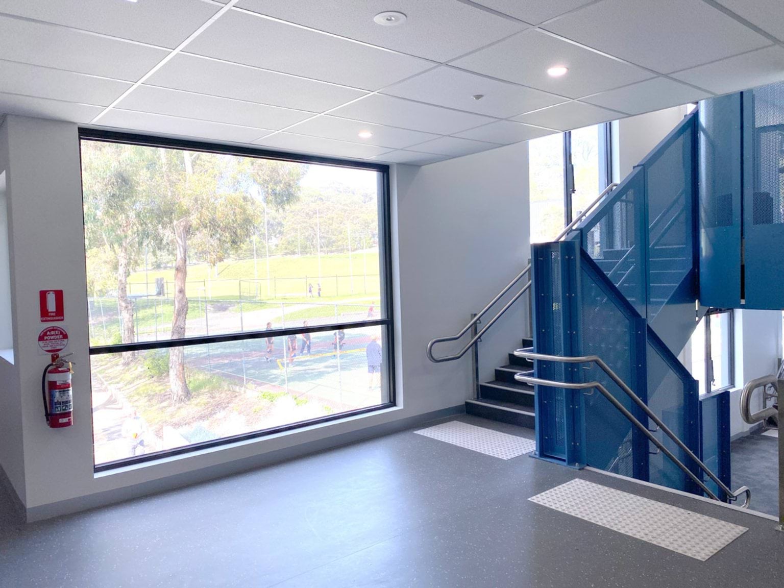 An interior staircase with blue metal panels and stainless steel handrails next to a large window overlooking outdoor sports courts.