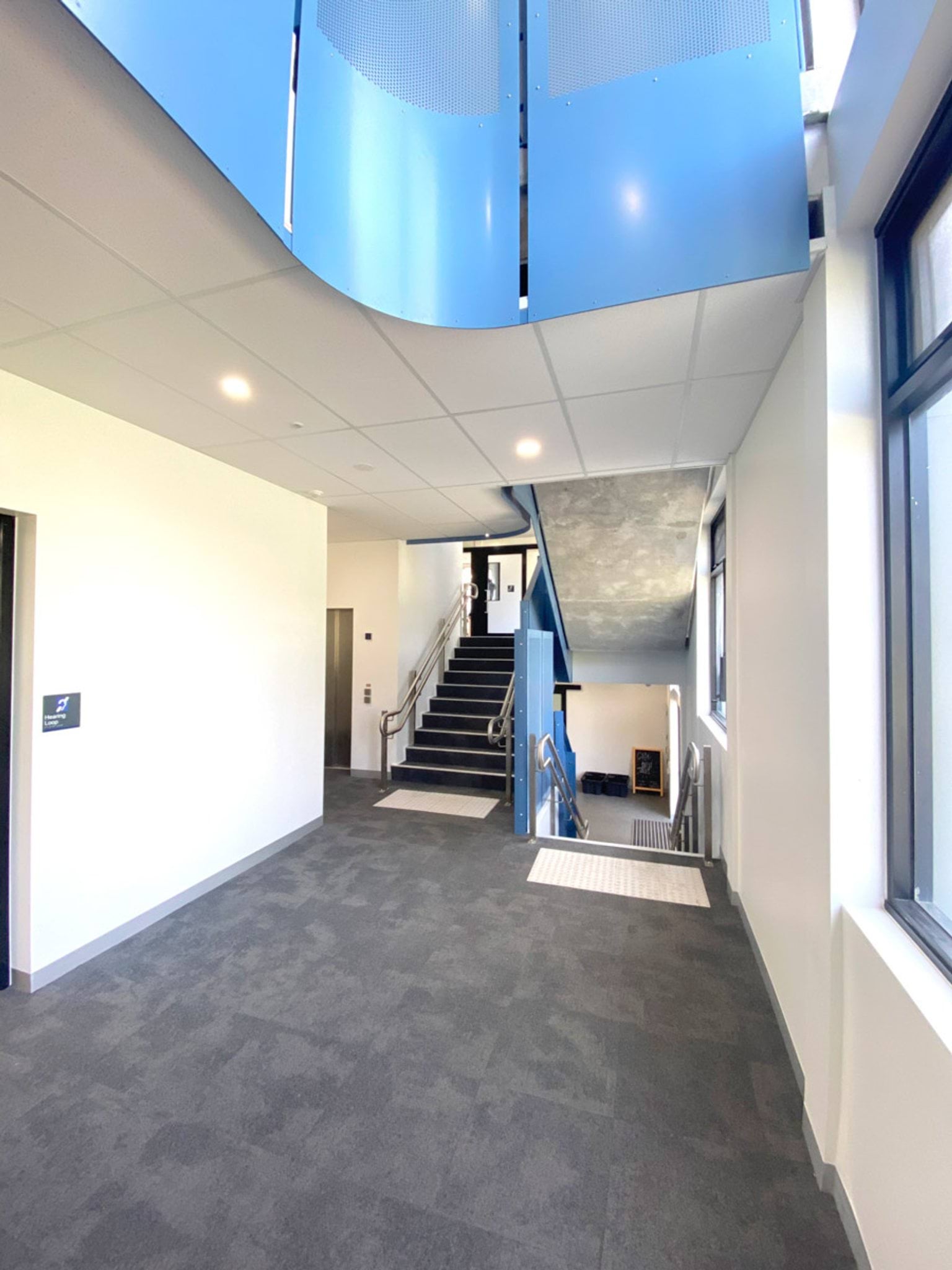 A stairwell with perforated blue metal panels and stainless steel handrails. Large windows allow natural light into the space.
