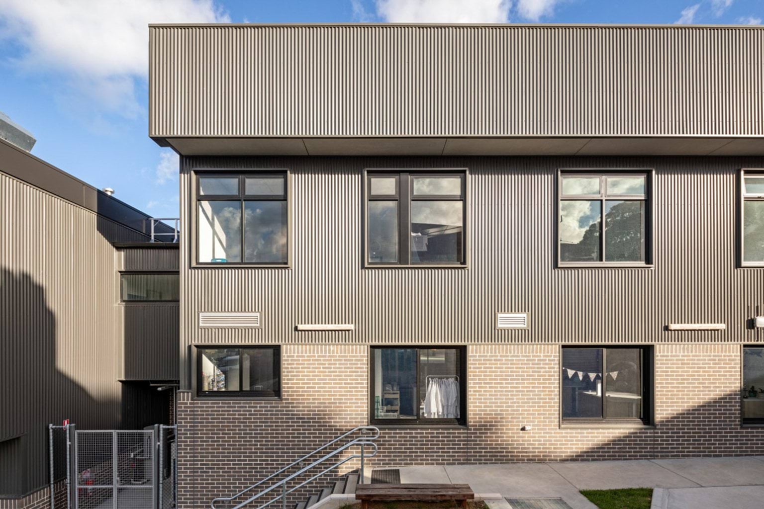 Front elevation of a school building with grey corrugated cladding, brickwork, and large windows. A concrete path and steps lead to the entrance.