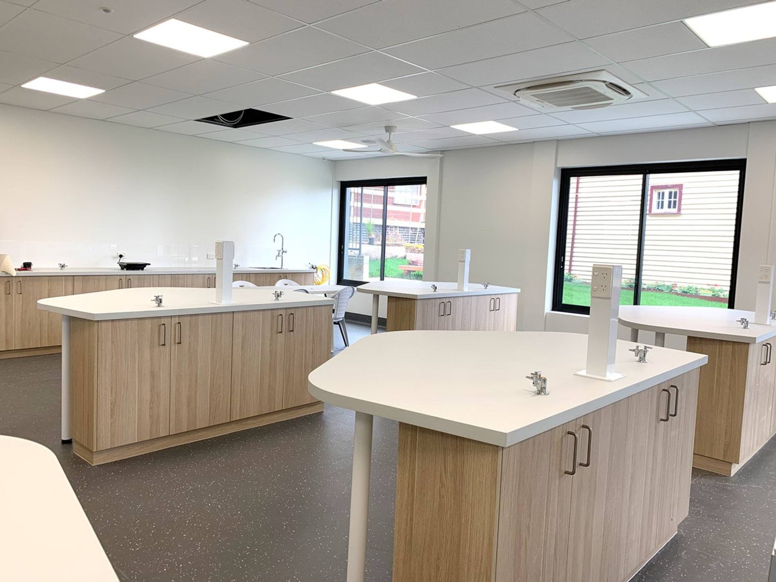 A science classroom with timber cabinetry, white benchtops, and integrated sinks. Large windows provide natural light and views of the outdoor area.