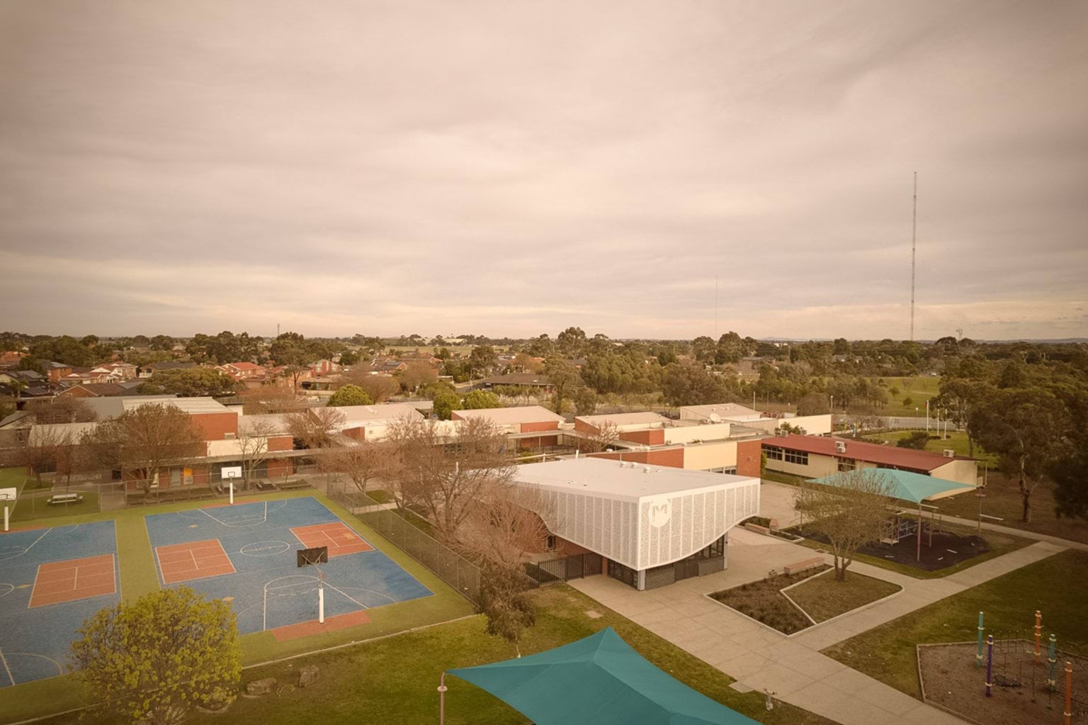 Aerial view of a school campus with multiple low-rise buildings, a sports court with blue and red markings, and landscaped paths surrounded by trees.