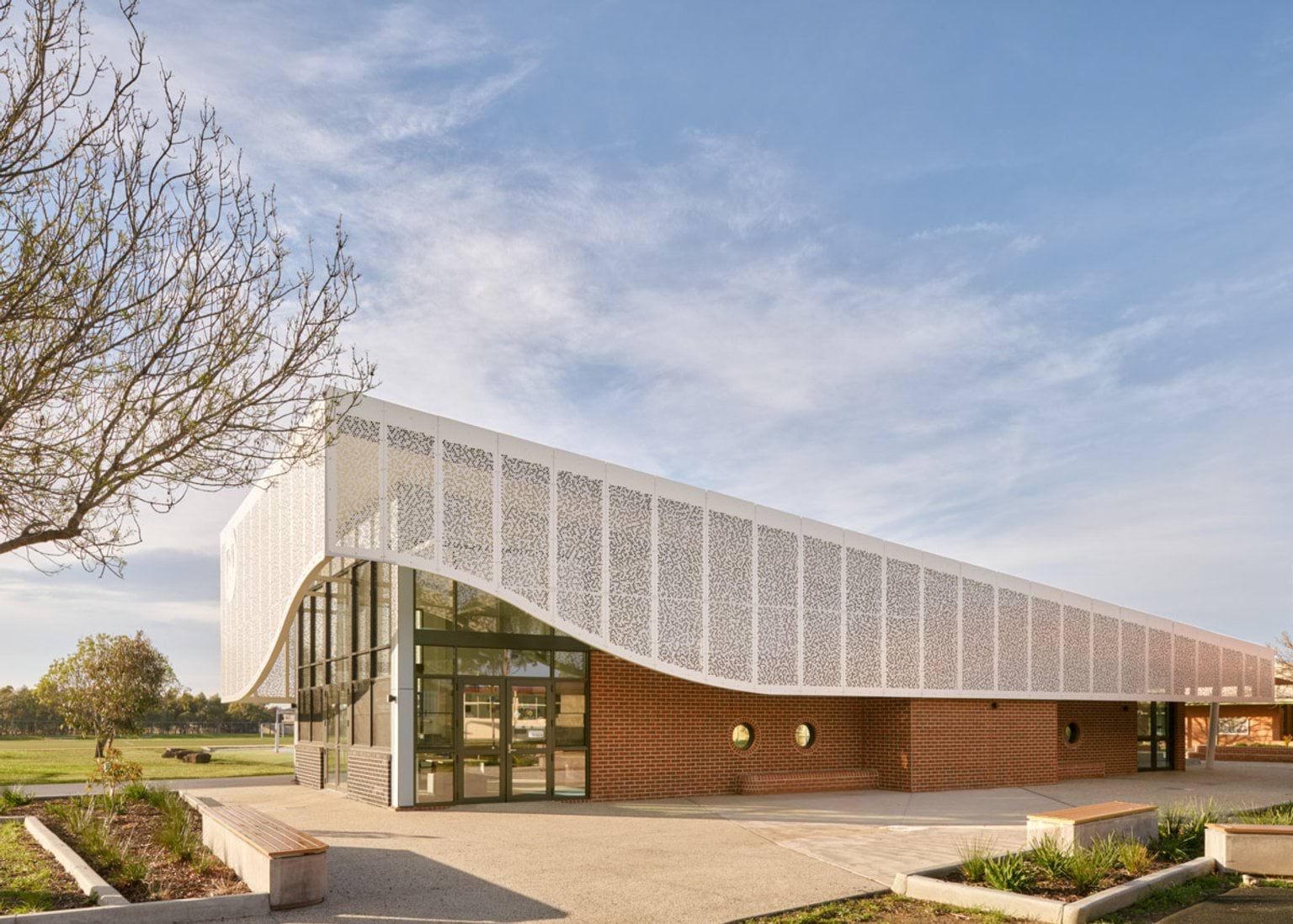 The exterior of a modern school building with a curved white patterned facade over red brick walls and large glass windows. Landscaping and seating are in the foreground.
