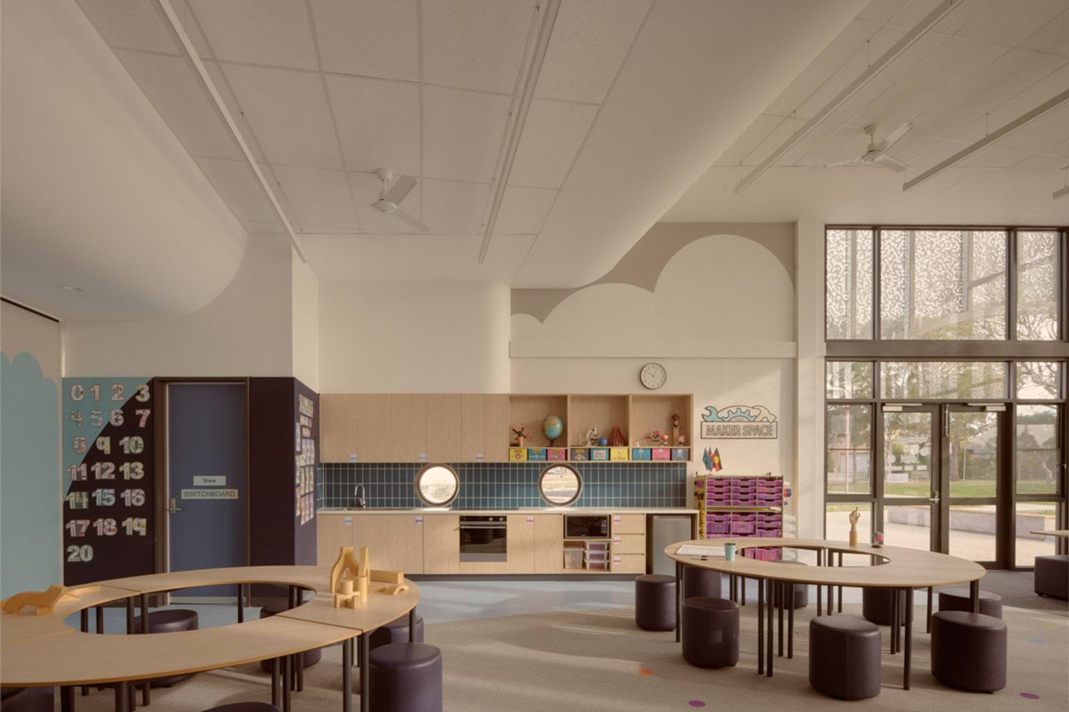 A maker space classroom with curved tables, round stools, and timber cabinetry. Two circular windows are set into a tiled splashback above a kitchenette.