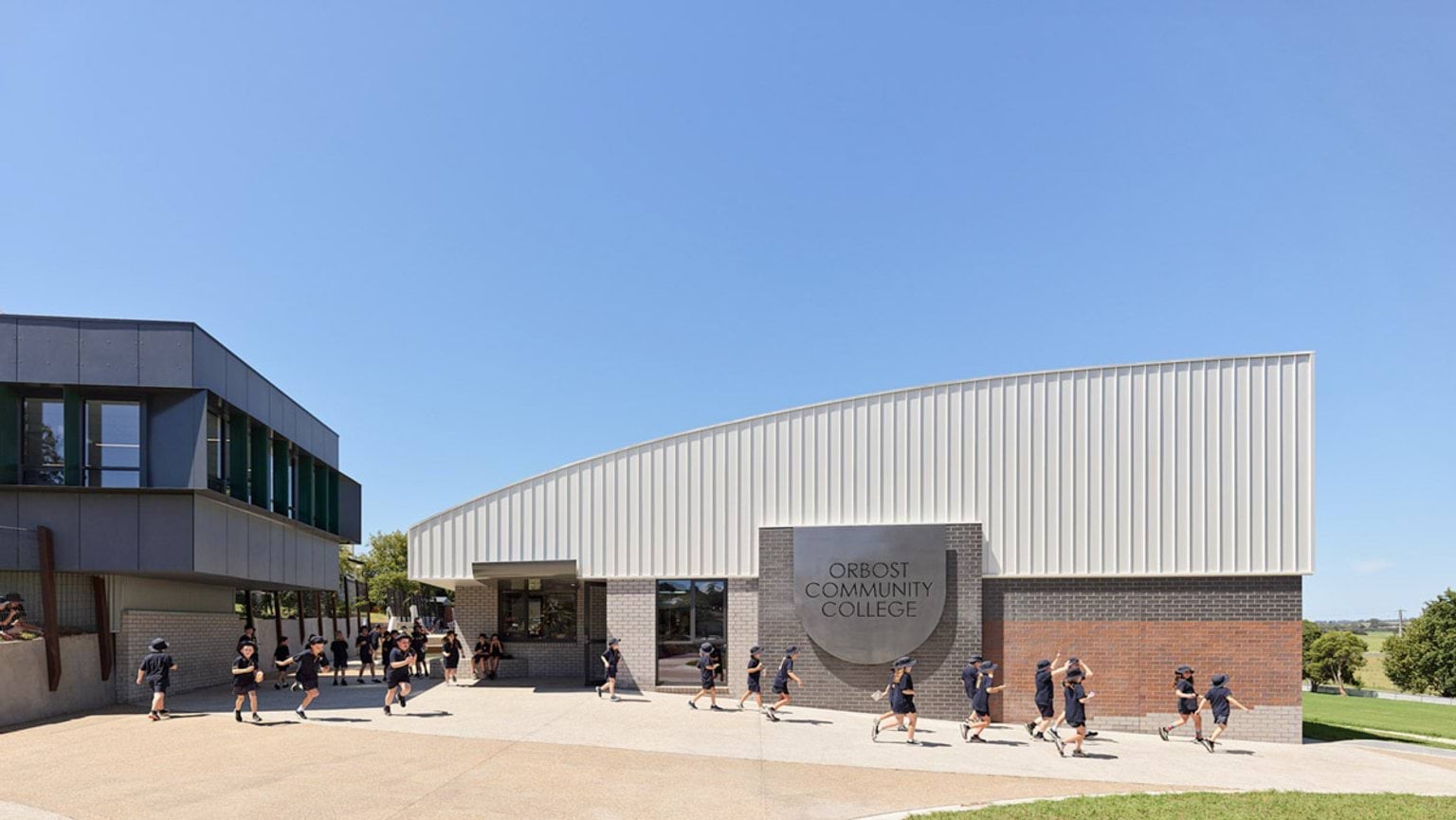 Modern school building with curved metal roof and brick facade, students walking across paved courtyard