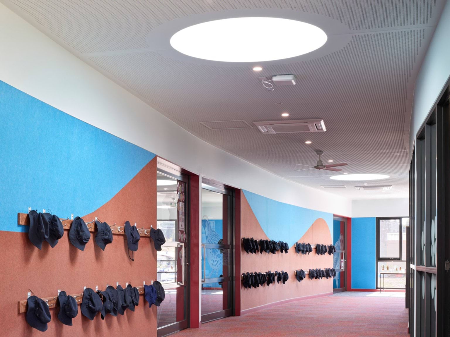 School corridor with colourful wave-patterned walls and rows of hooks holding hats, circular skylight above