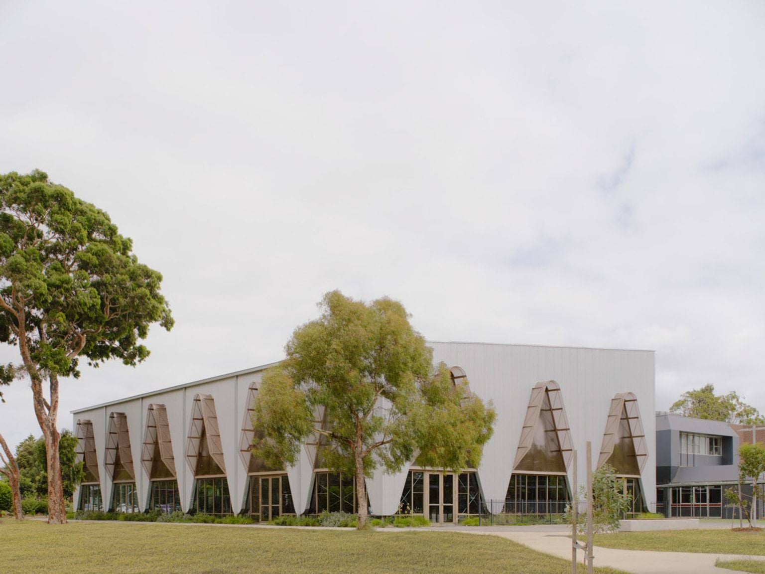A modern school building with a white facade and triangular feature panels over glass windows. A paved path and landscaped garden beds lead to the entrance, with trees framing the view.