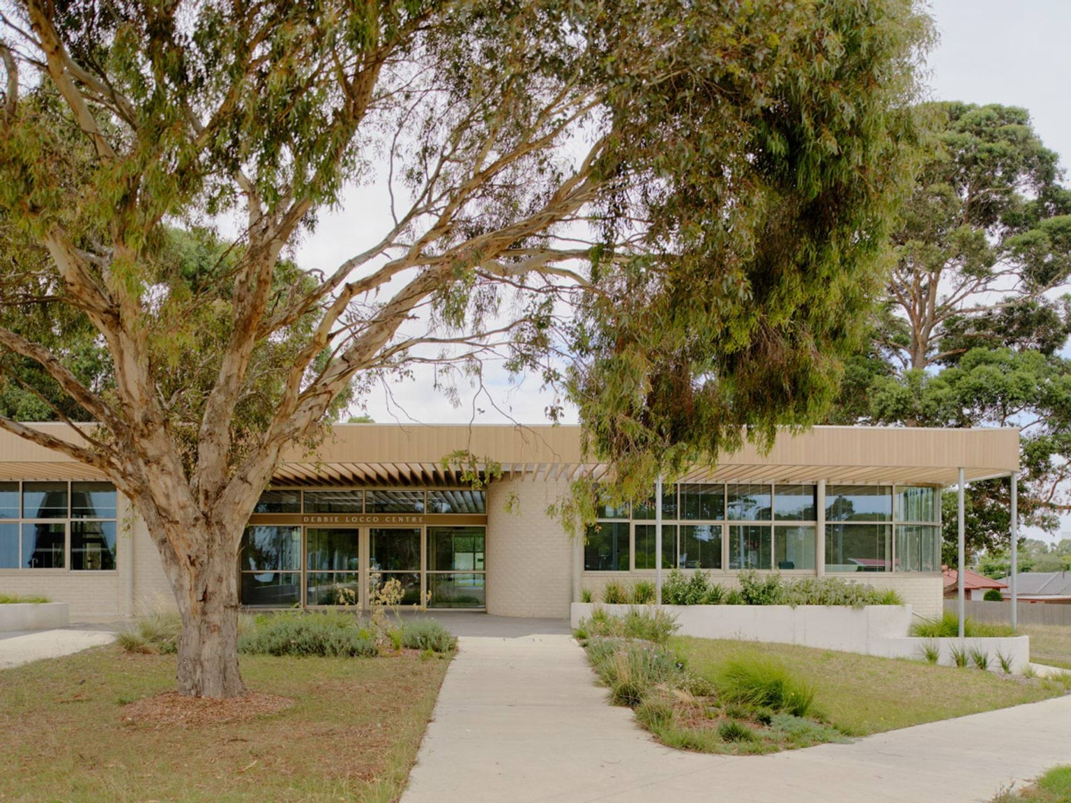 A modern school building with a timber facade and large windows, surrounded by trees and landscaped garden beds. An open classroom is visible through the glass doors.