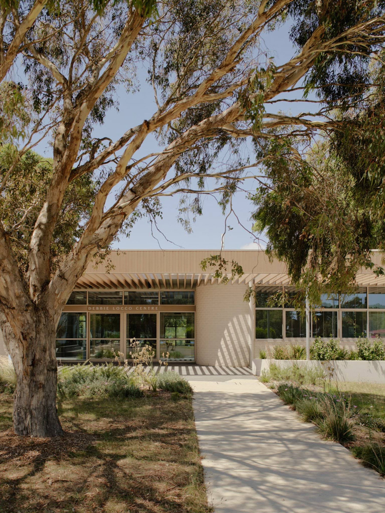 The entrance to the centre with a paved path leading to glass doors. The building has a light timber facade and landscaped garden beds under mature trees.