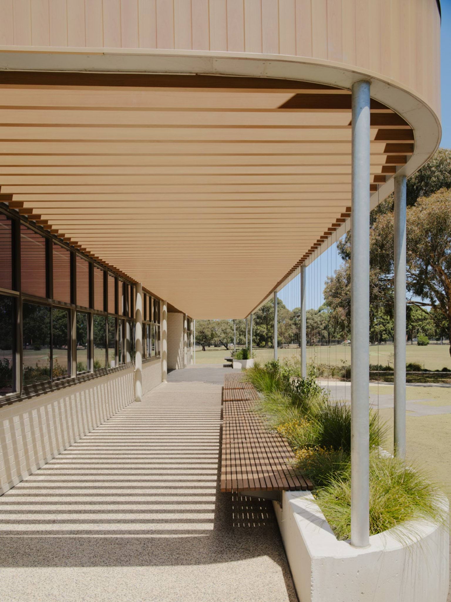 A covered outdoor walkway with timber bench seating and landscaped garden beds. Vertical posts support the roof, creating striped shadows on the paved surface.