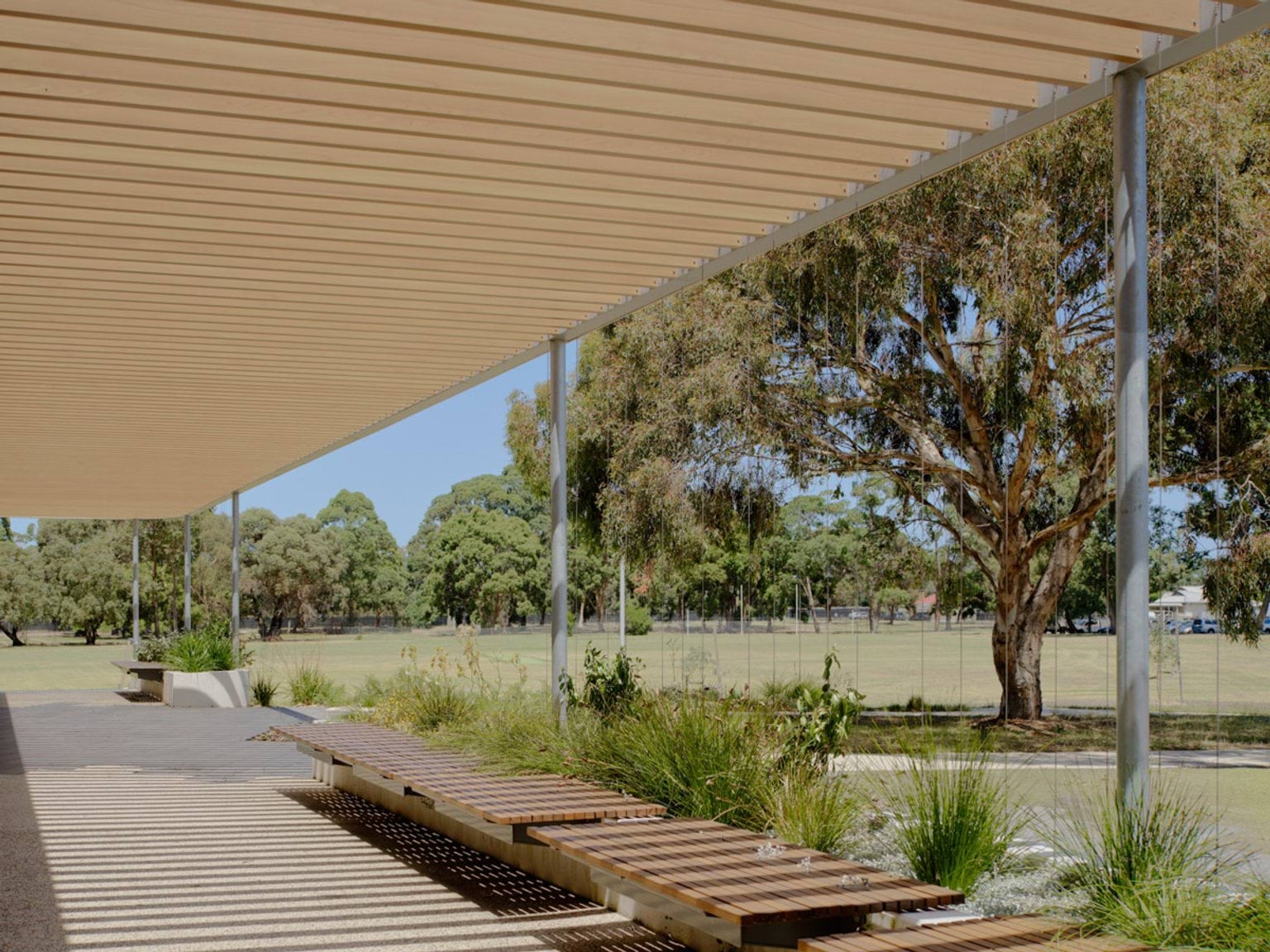 A covered outdoor seating area with timber benches and landscaped garden beds, overlooking a grassed sports field and mature trees.