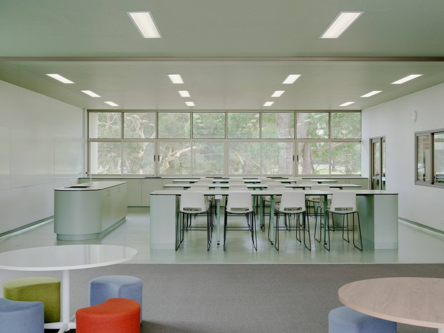 A bright science classroom with long lab benches and white stools arranged in rows. Large windows provide views of trees outside, and the space has clean white walls and ceiling lights.