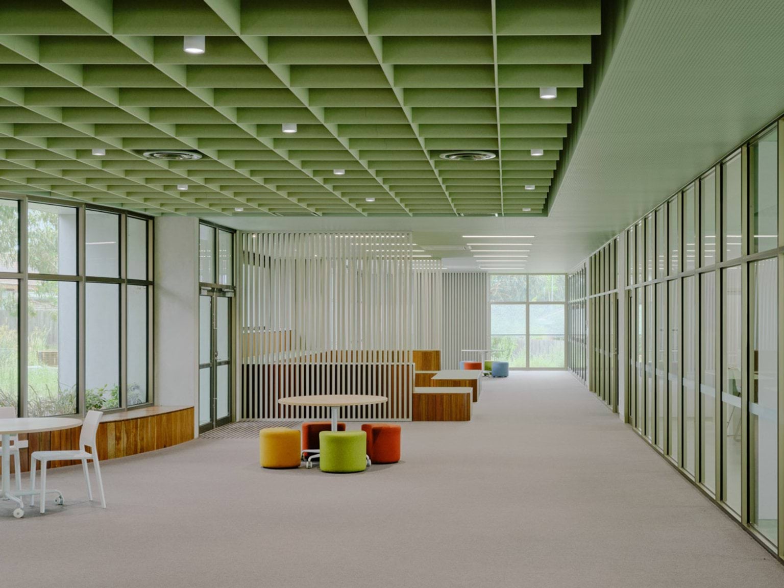 An open learning space with a green grid ceiling, glass walls, and soft seating in bright colours. Timber benches and tables are arranged throughout the area.