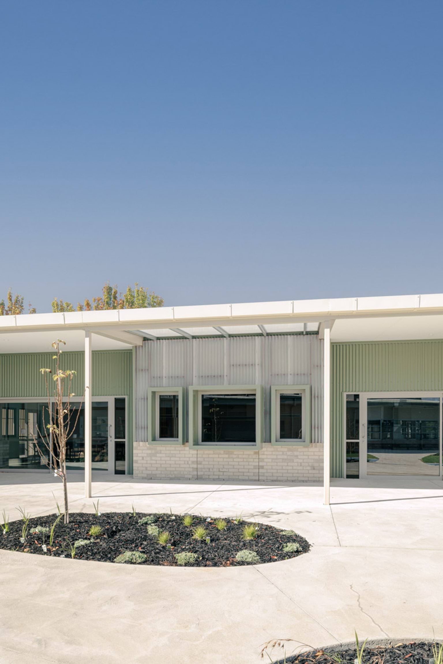 The exterior of a school building with green and cream cladding, light brickwork, and a covered walkway. A circular garden bed with young plants is in the foreground.