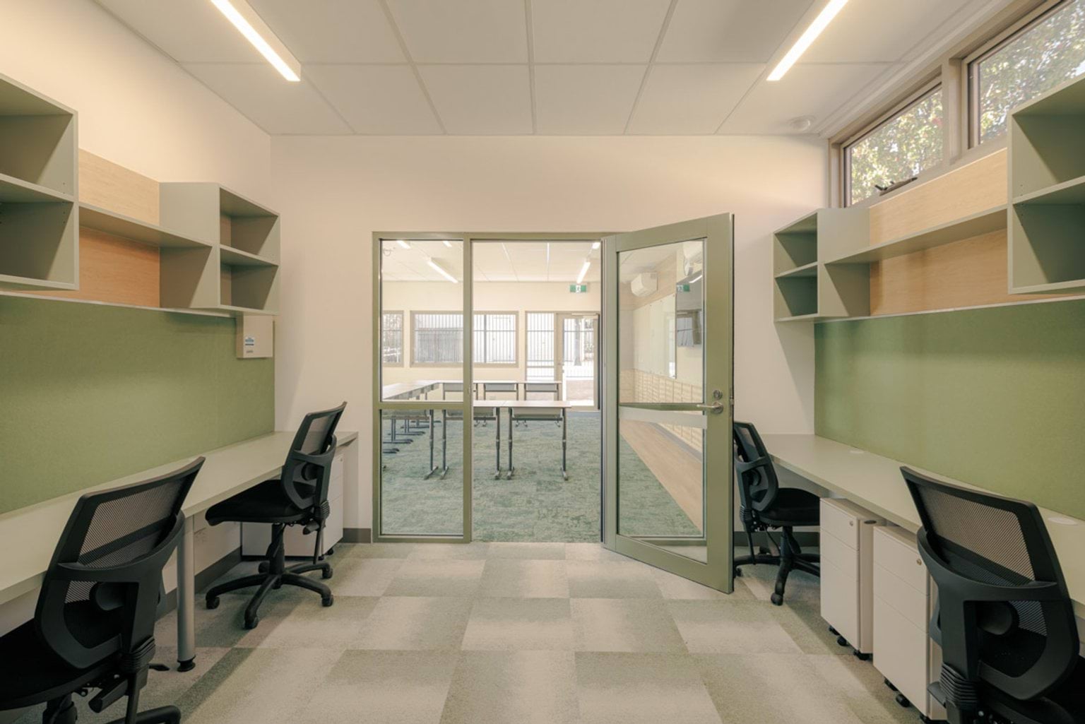An office with two workstations, green wall panels, and shelving. A glass door opens to a classroom with tables and chairs.