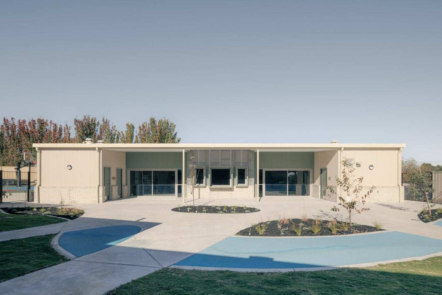 The front of a modern school building with green and cream cladding, light brickwork, and a covered entrance. Landscaped garden beds and curved paths are in the foreground.