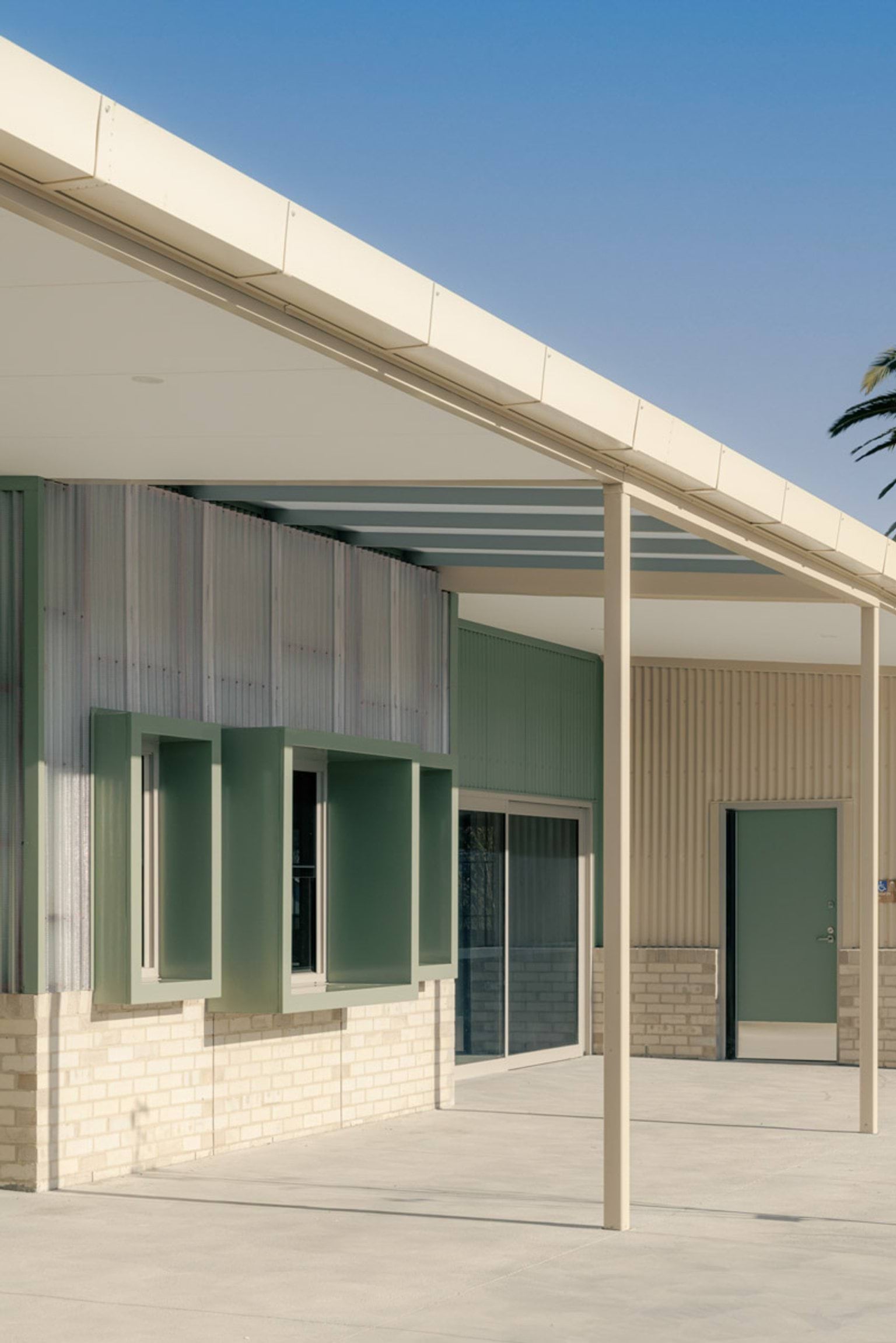School exterior with a wide covered walkway, green box-shaped recesses, and corrugated cladding above light brick walls.