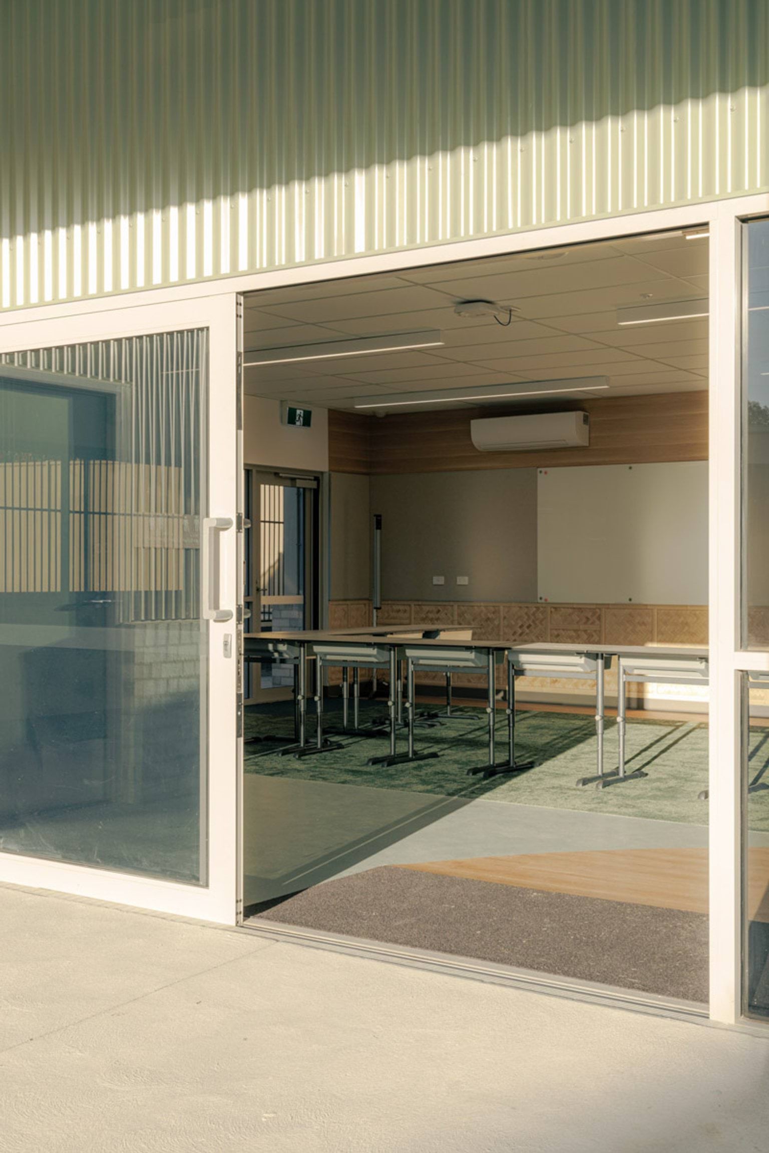 Exterior view through sliding glass doors into a classroom with tables and chairs on green carpet. The building has timber and light-coloured walls.