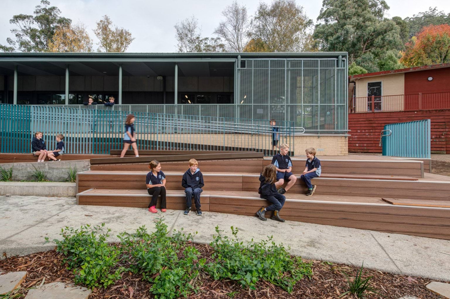 Students sitting and walking on timber tiered seating in a landscaped outdoor area with garden beds. A modern school building with teal fencing and large windows is in the background.