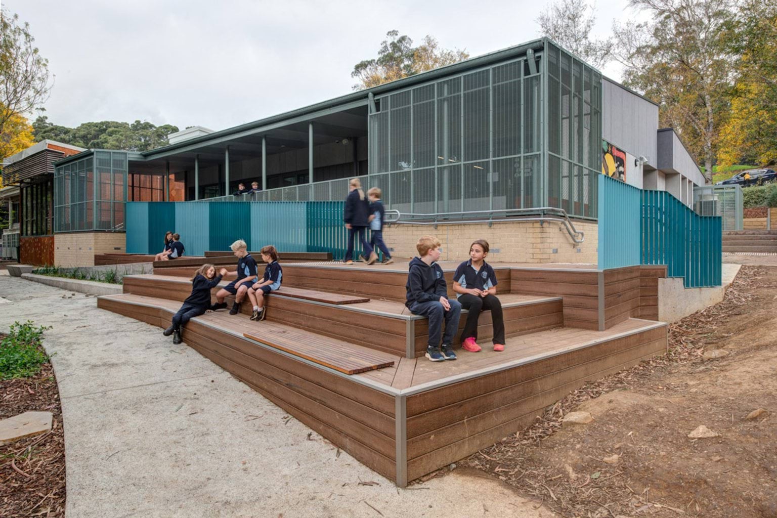 Students sitting and walking on timber tiered seating in front of a school building with teal fencing and large windows. Landscaping surrounds the seating area.