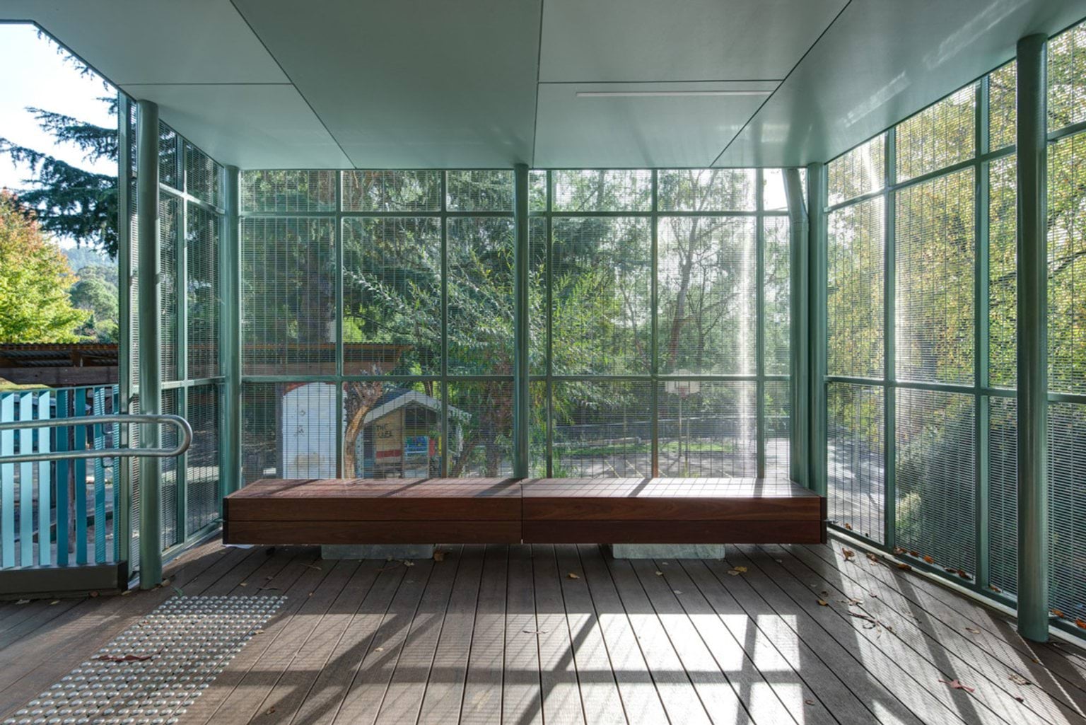 A covered outdoor area with timber decking and built-in timber bench seating, enclosed by green metal mesh panels with views of surrounding trees.