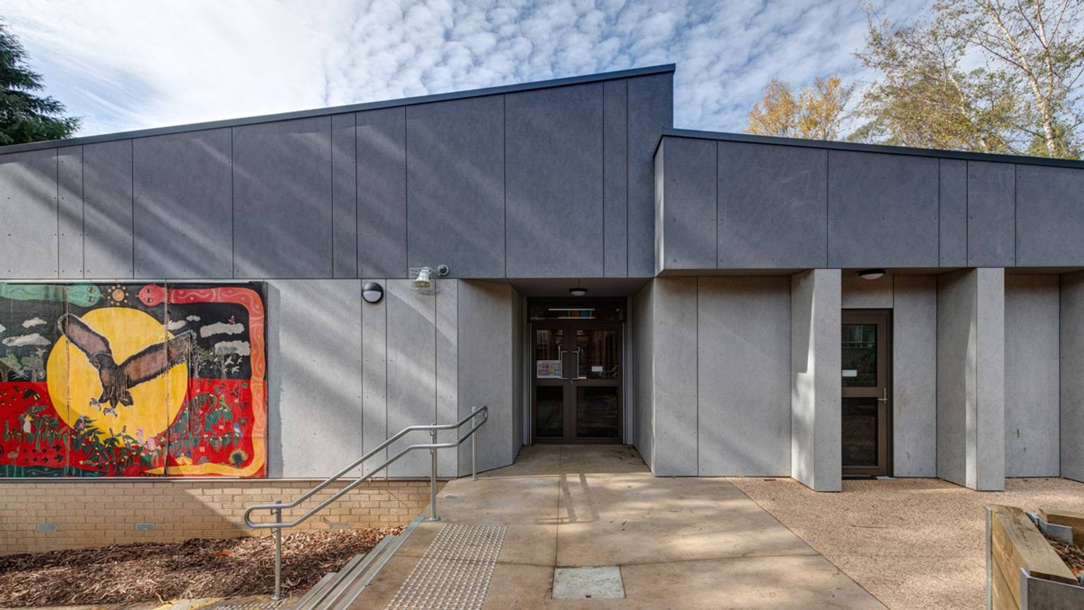 The entrance of a school building with grey cladding and a colourful mural featuring a bird and a yellow sun. A ramp with metal handrails leads to the doorway.