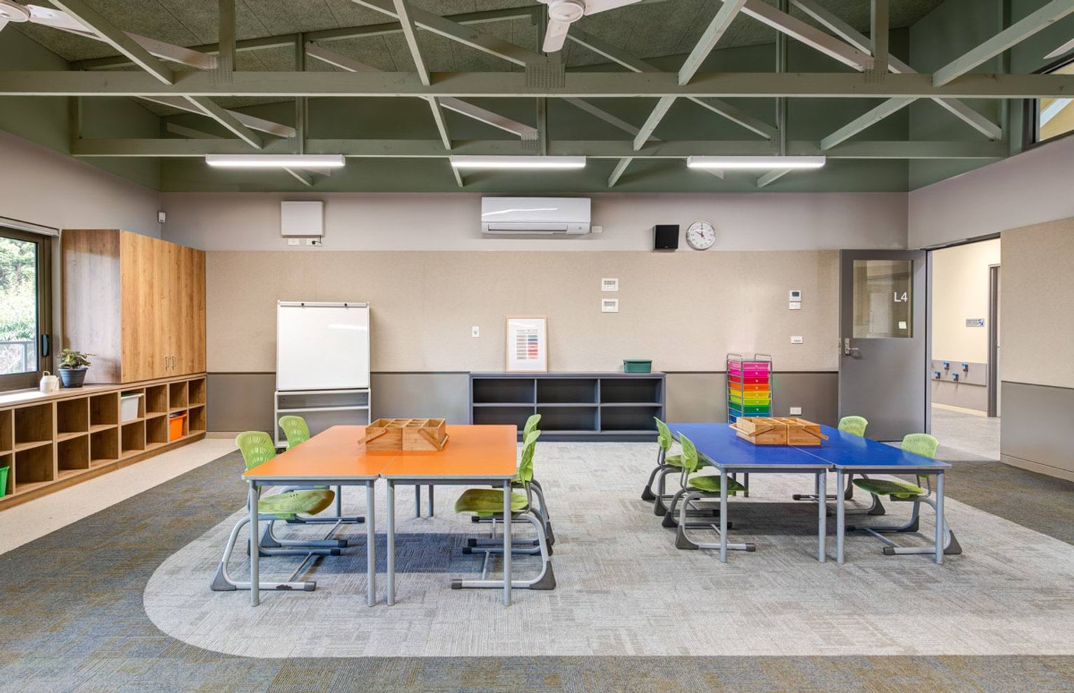 A classroom with orange and blue tables, green chairs, and timber storage units. The ceiling has exposed beams and LED lights.