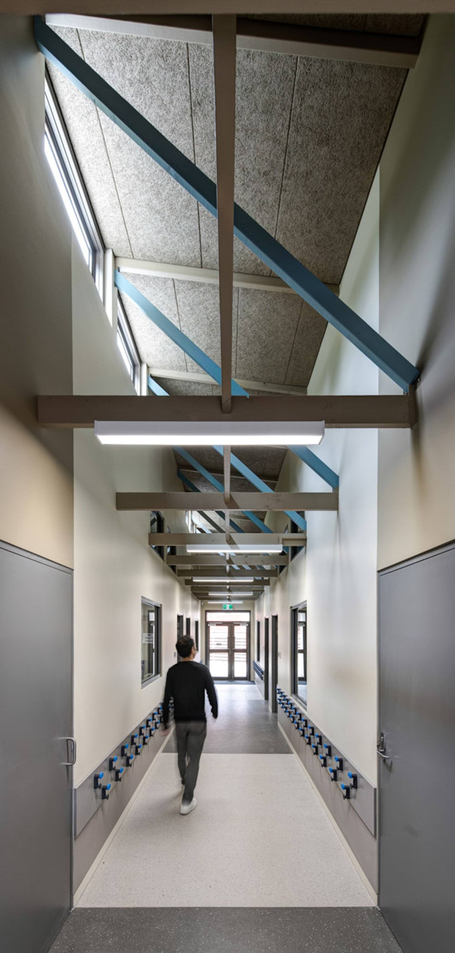 A long hallway with high ceilings and exposed blue beams. Hooks line the walls beneath windows, and a person is walking toward the exit.
