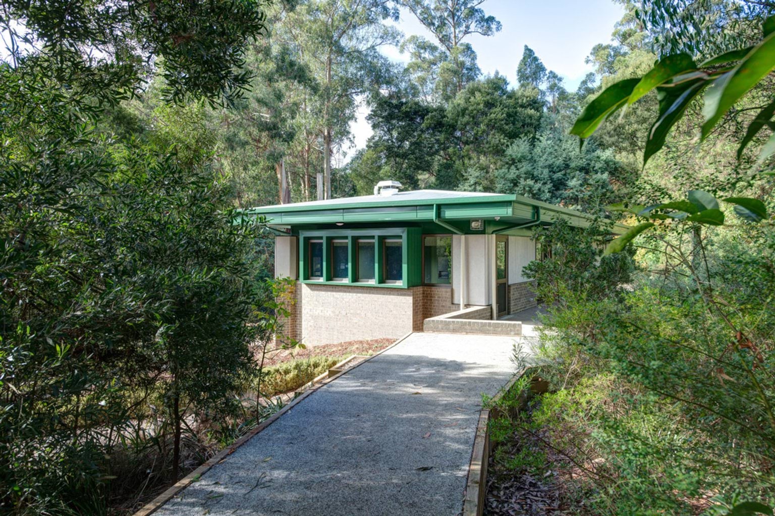 A concrete pathway leading to a school building with a green roof and brick walls, surrounded by dense bushland and landscaped garden beds.