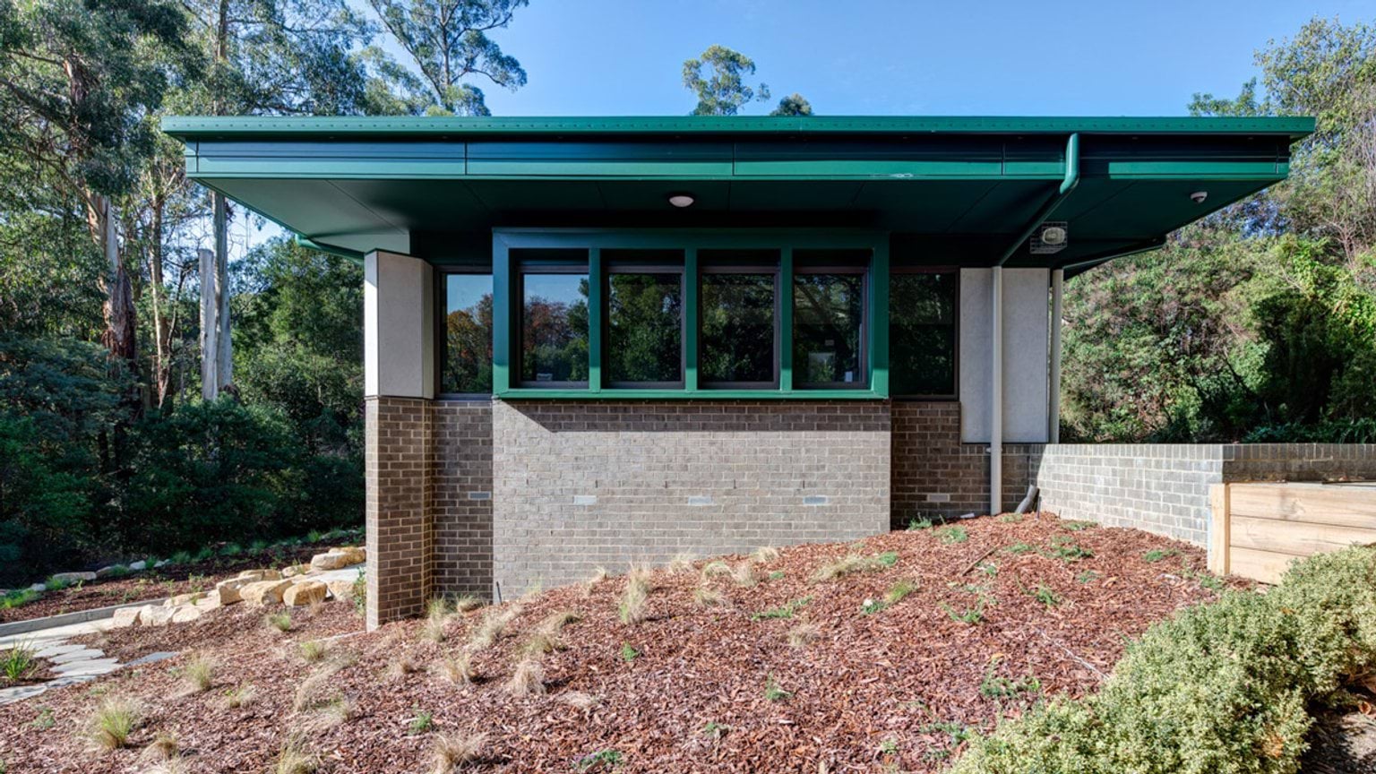The exterior of a school building with a green roof, brick walls, and landscaped garden beds surrounded by bushland.