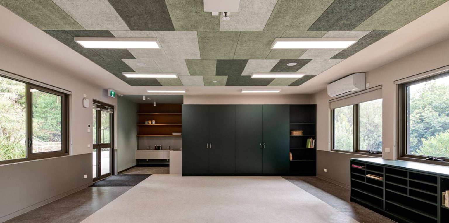A bright learning space with patterned green acoustic ceiling panels, large windows, and dark green cabinetry. A small kitchenette is visible in the corner.