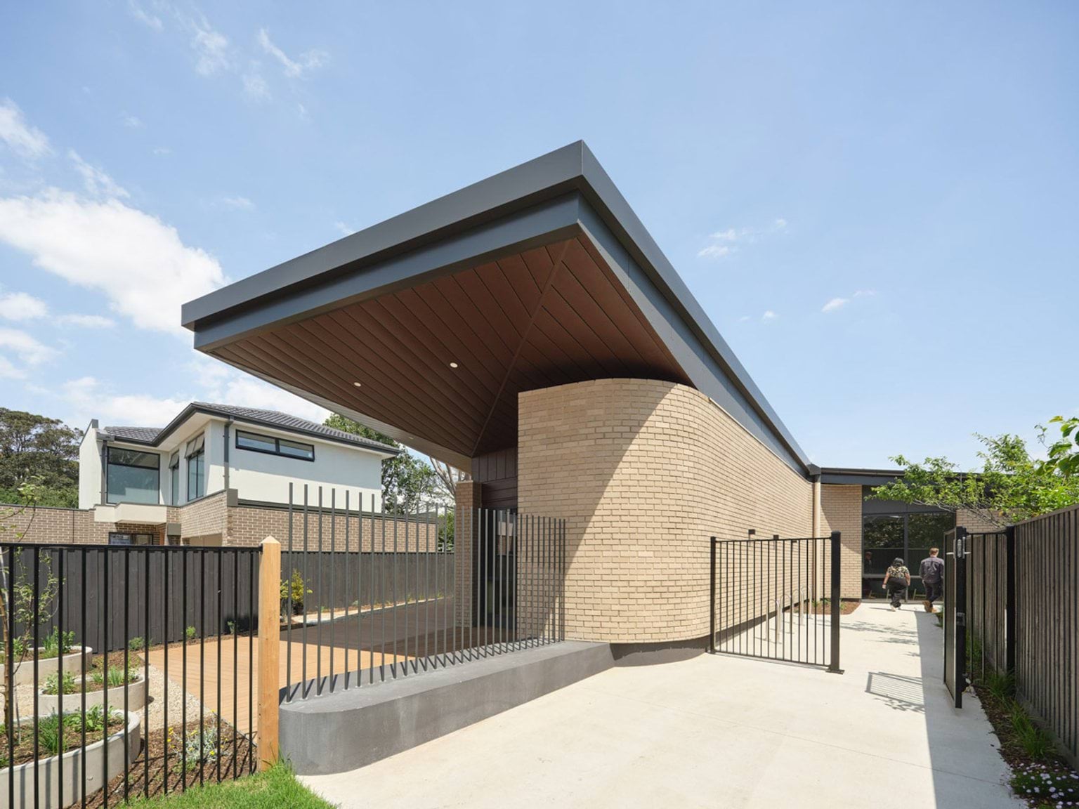 The exterior of the kindergarten with cream brick walls and a bold curved roof in dark grey. A black metal fence encloses the entrance, and landscaped garden beds line the path.
