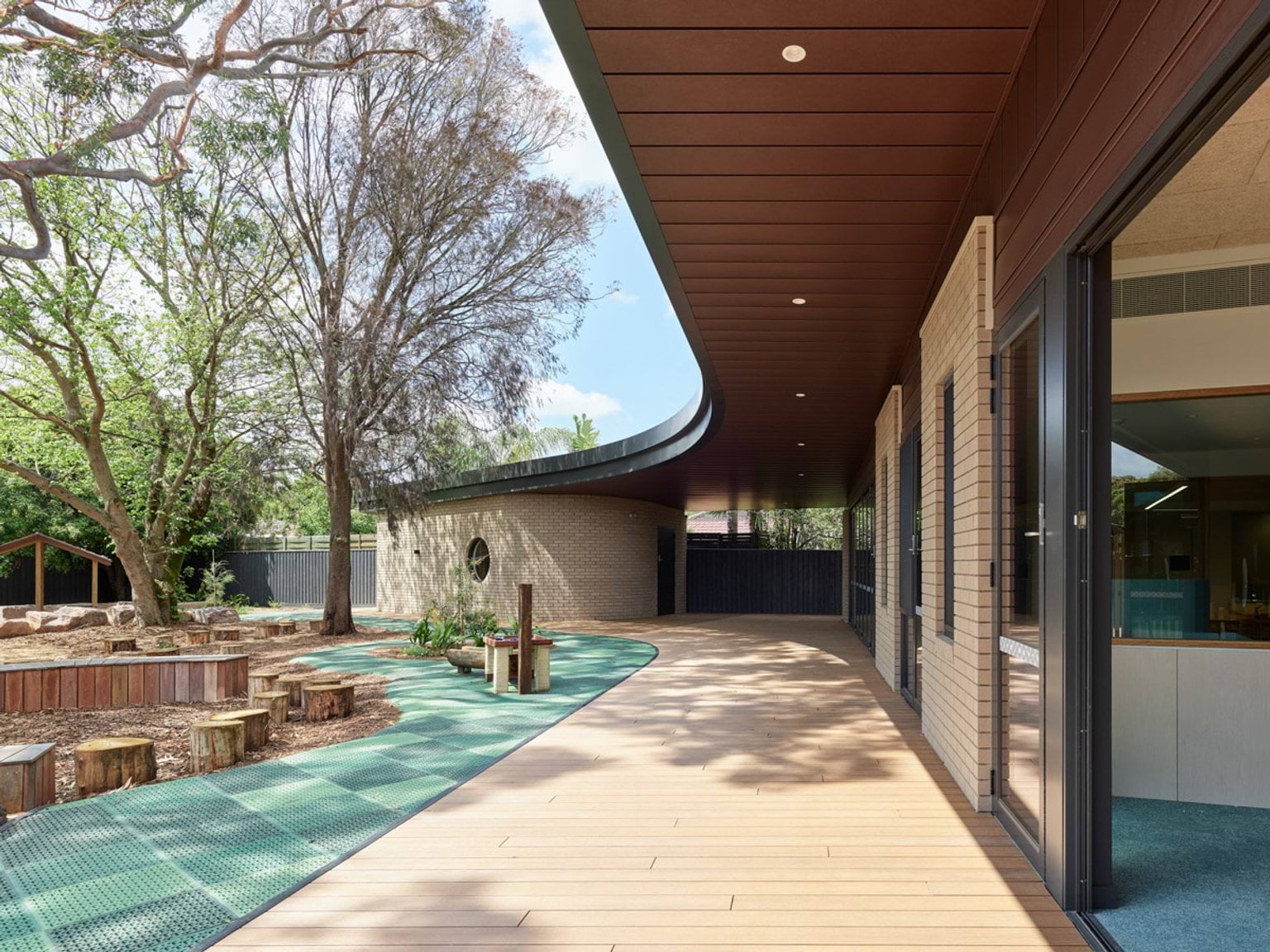 A curved section of the kindergarten exterior with cream brick walls and a round window. Timber decking and green play mats lead to a black fence.