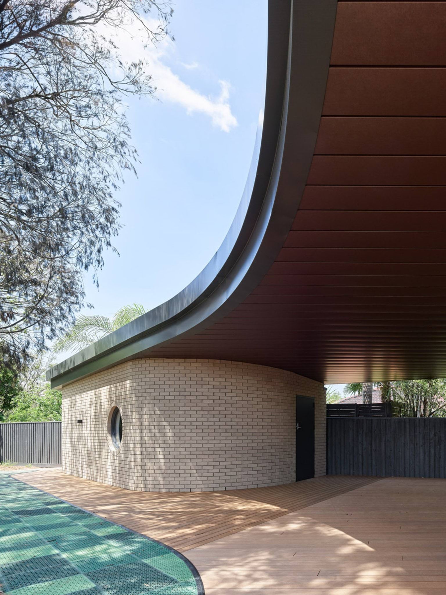 The kindergarten outdoor play area with timber seating, garden beds and a large tree. The curved building has cream brick walls and a round window.