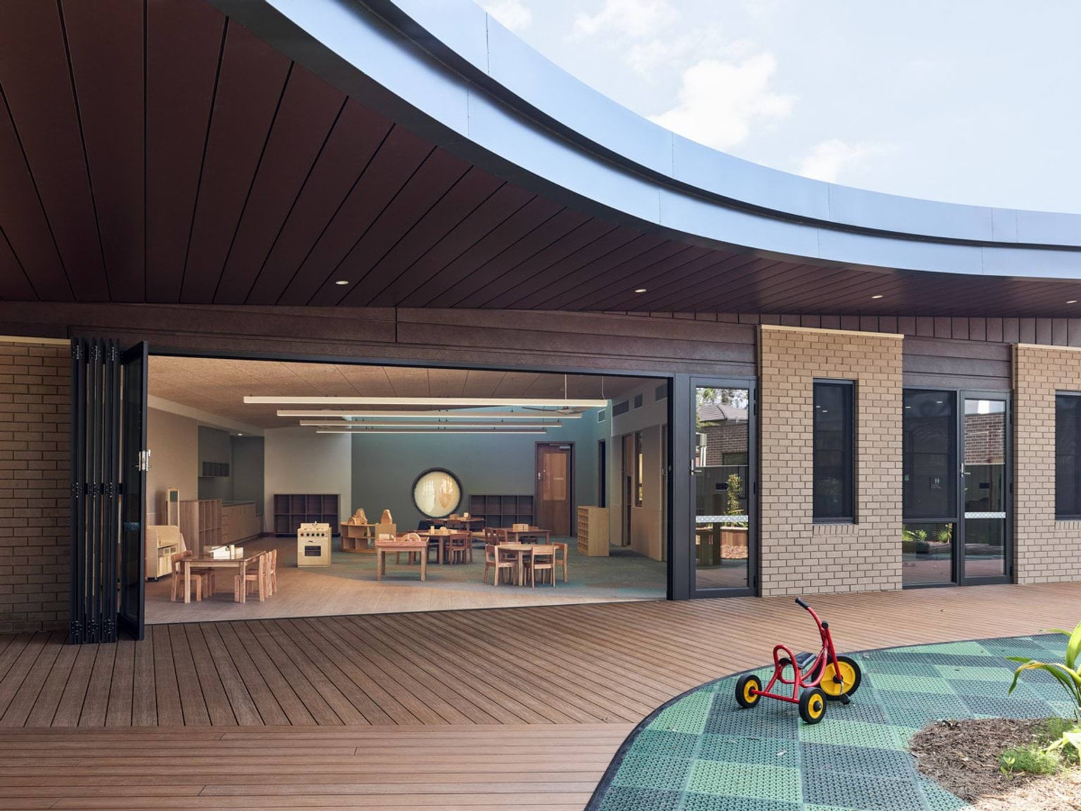A kindergarten classroom with timber tables and chairs, pale walls and a cork-panel ceiling. Large glass doors open to the outdoor play area with trees and garden beds.