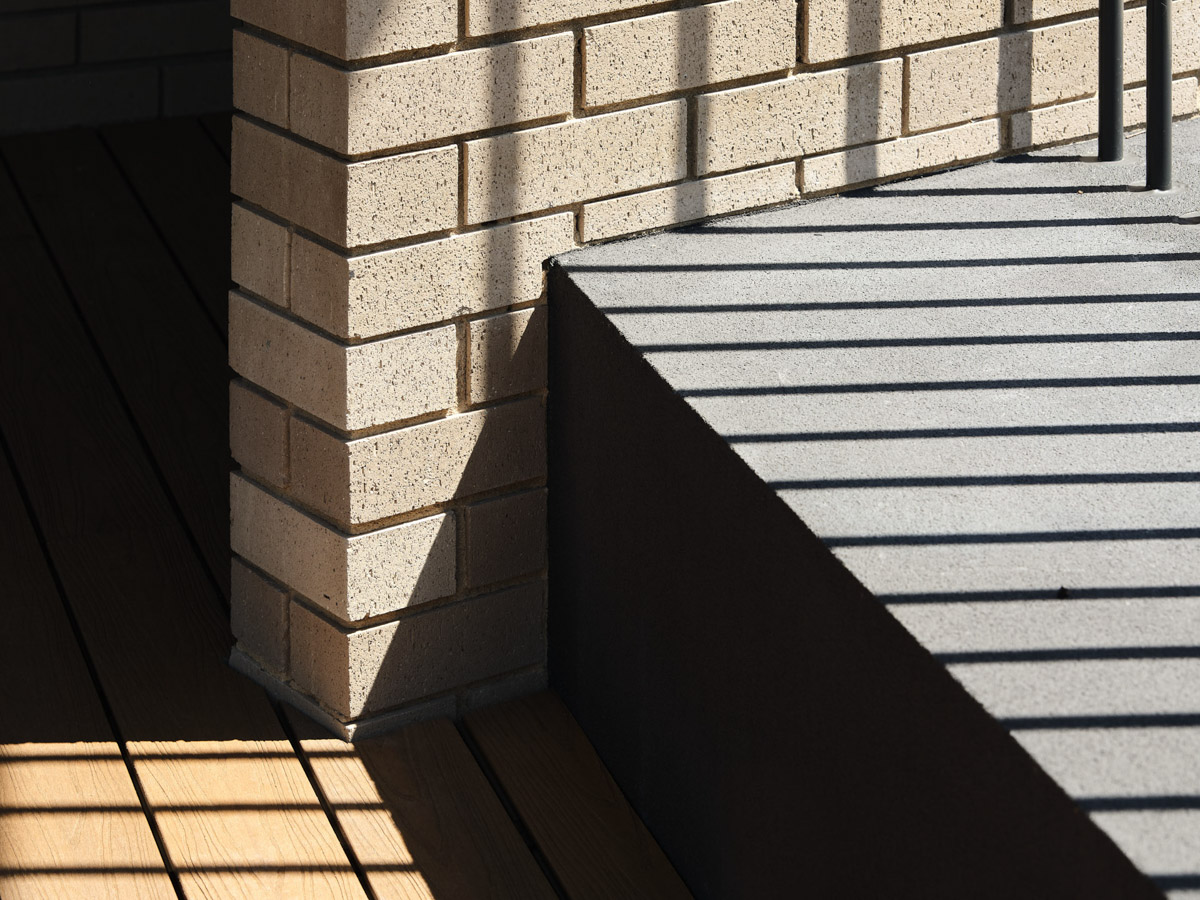 The outdoor play area with timber decking, green play mats and garden beds. The kindergarten building curves around the space with cream brick walls and a dark roof.