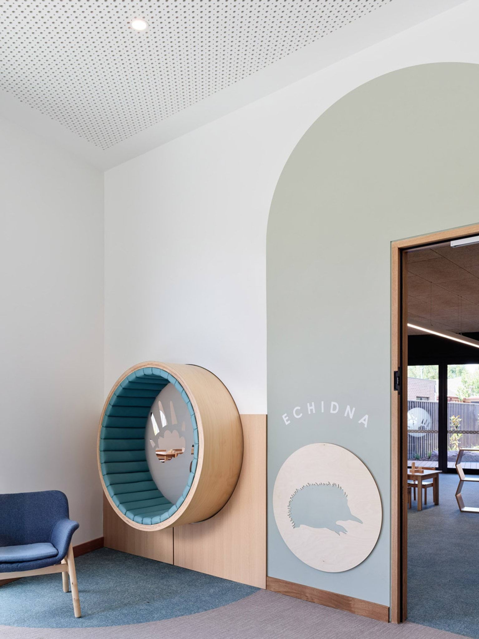 A circular timber seating nook built into a wall, with soft blue cushions and an animal footprint design. A blue chair sits nearby and a sign reads “Echidna”.