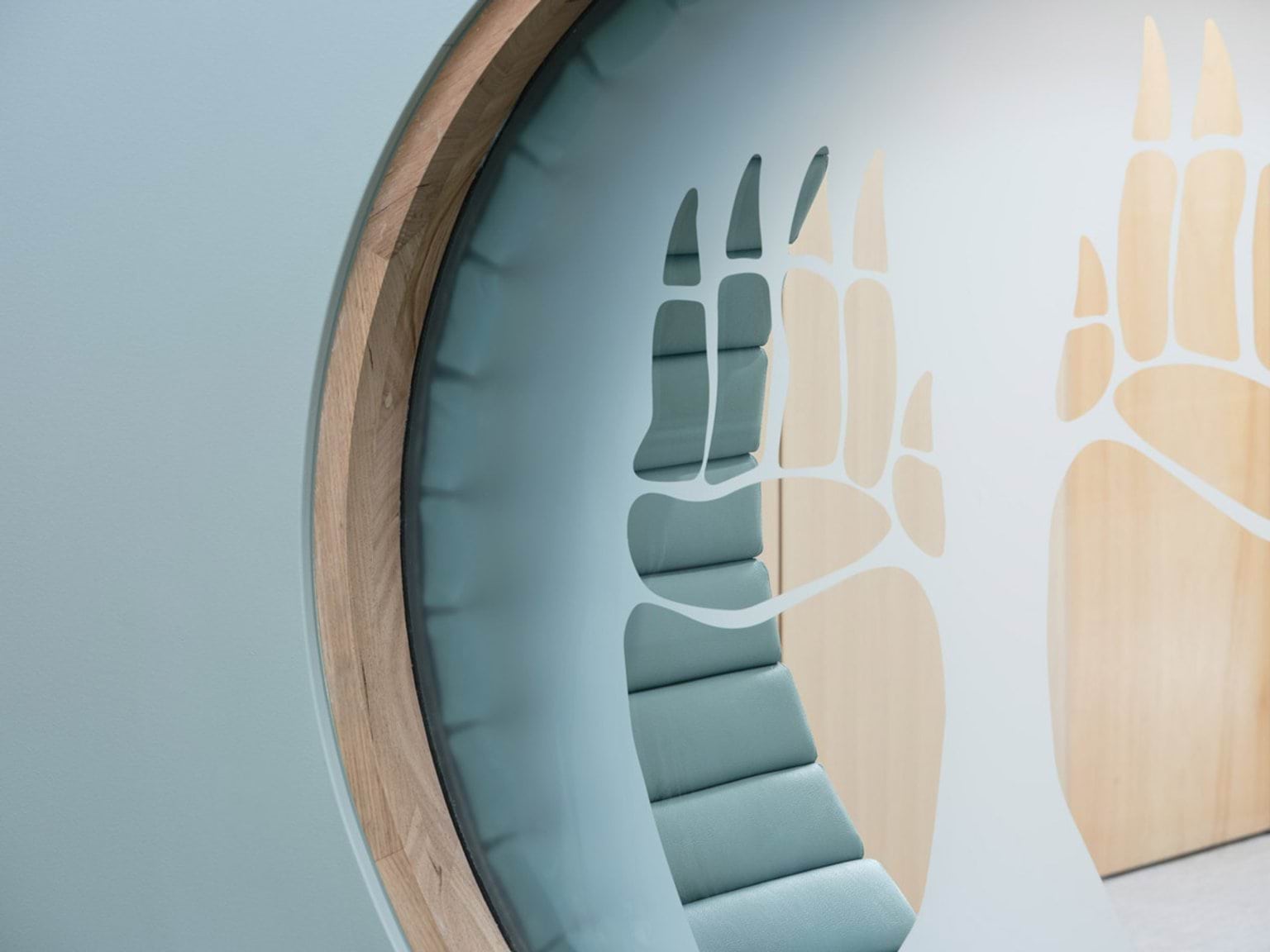 A circular timber seating nook built into a pale green wall, with soft blue cushions. The cut-out design features stylised animal footprints.