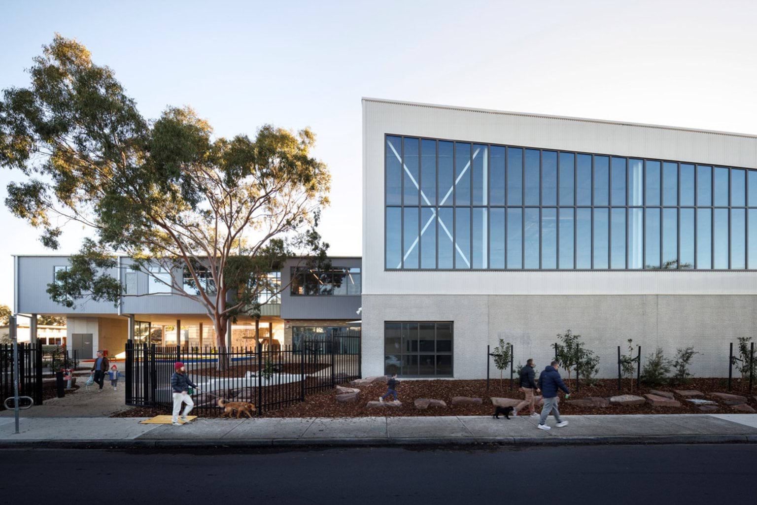The exterior of a modern school building with tall glass windows, landscaped garden beds, and a large tree in front. A black metal fence surrounds the entrance.