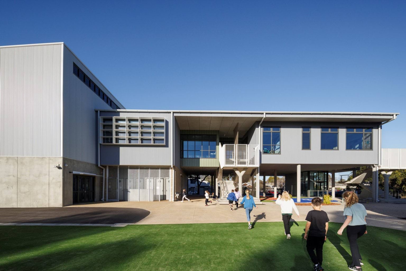 Students walking across a green outdoor play area towards a two-storey school building with wide windows and an open undercroft.